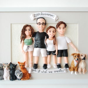 May include: A framed 3D clay sculpture of a family with two dogs and four cats. The family is made up of two adults and two children. The adults are holding hands and the children are standing in front of them. The sculpture is on a white background and has the text "20th Anniversary!" and "look at what we created!" written on it.