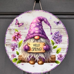 May include: A purple gnome holding a sign that says "Hello Spring!" surrounded by purple flowers and butterflies on a white wooden background.