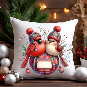 May include: White throw pillow with a red cardinal and a brown bird wearing winter hats and perched on a red and black plaid ornament. The ornament has a blank name tag.