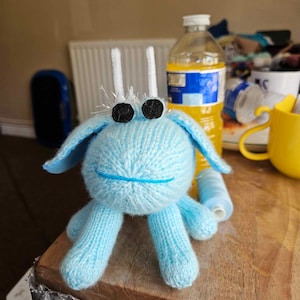 Puede incluir: Una criatura tejida de color azul claro con ojos negros, antenas blancas y una boca cosida azul. El juguete hecho a mano tiene cuatro patas y dos orejas. La criatura está sobre una superficie de madera.
