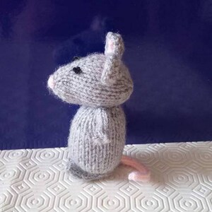 May include: A gray knitted mouse toy with pink ears and a black nose. The mouse is standing on a white surface with a hexagonal pattern.