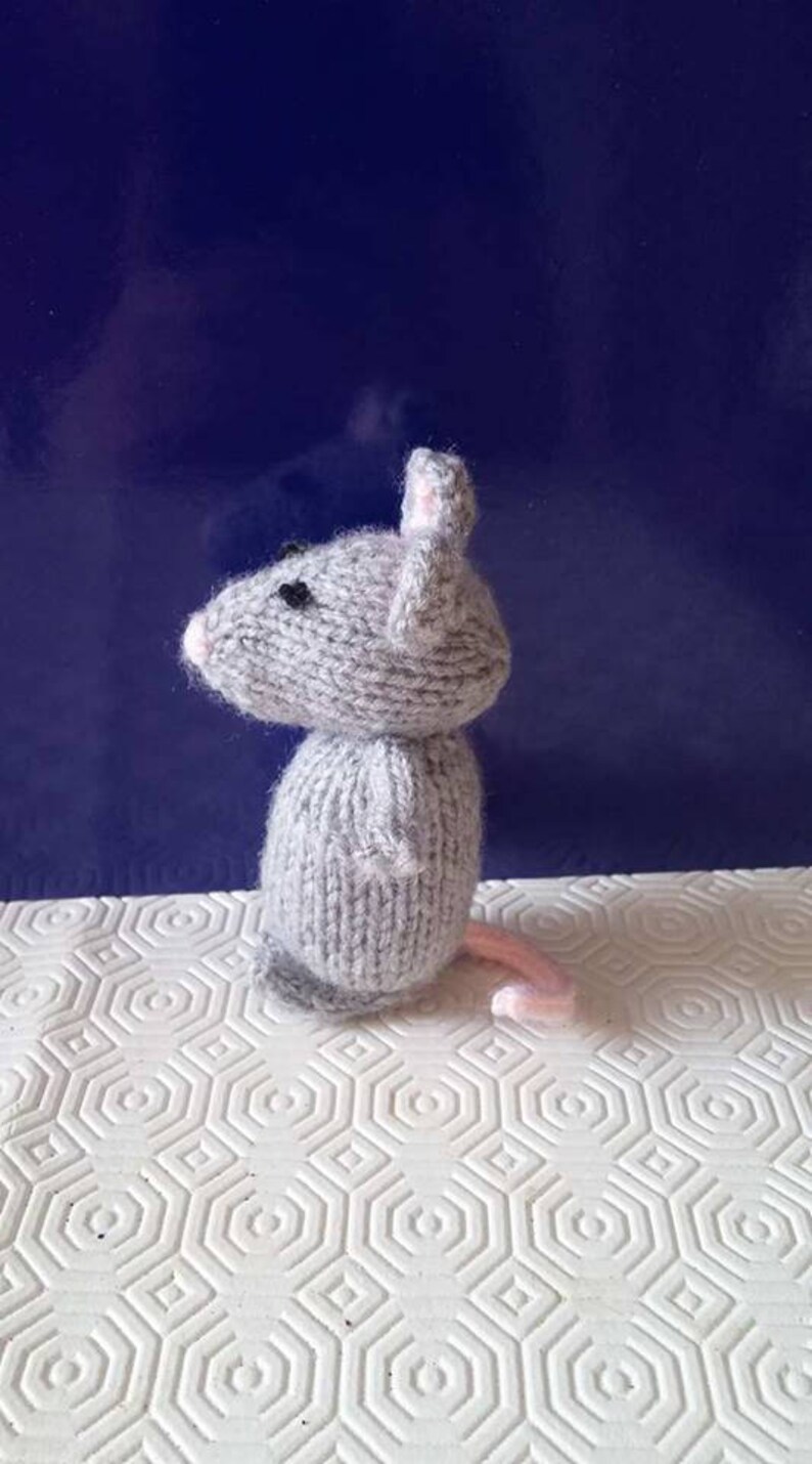 May include: A gray knitted mouse toy with pink ears and a black nose. The mouse is standing on a white surface with a hexagonal pattern.