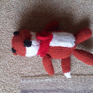 May include: A knitted fox toy with a red scarf. The fox is lying on its back with its legs and tail outstretched. The toy is made of orange, white, and black yarn.