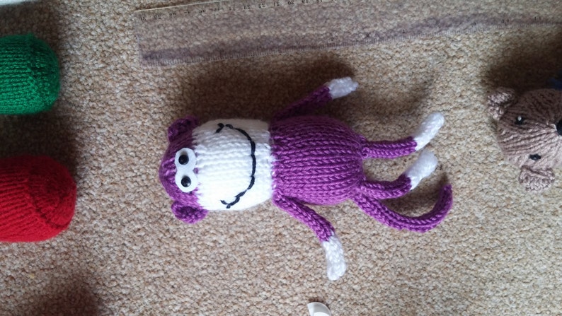 May include: A purple knitted monkey toy with white yarn for the face, arms, and legs. The monkey has black eyes and a white smile.