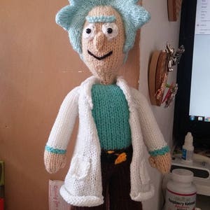 Könnte beinhalten: Ein gestricktes Plüschtier eines Charakters mit blauen Haaren, das einen weißen Laborkittel, eine braune Hose und schwarze Schuhe trägt. Das Spielzeug steht auf einem Tisch mit anderen Gegenständen.