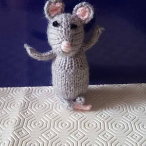 May include: A gray knitted mouse toy with pink ears and a pink nose. The mouse is standing on a white surface with a hexagonal pattern.