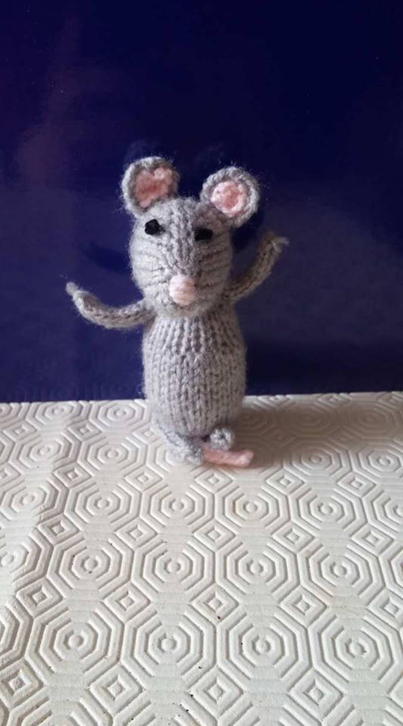 May include: A gray knitted mouse toy with pink ears and a pink nose. The mouse is standing on a white surface with a hexagonal pattern.
