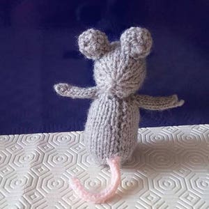 May include: A gray knitted mouse toy with pink ears and a pink tail. The mouse is sitting on a white surface with a hexagonal pattern.