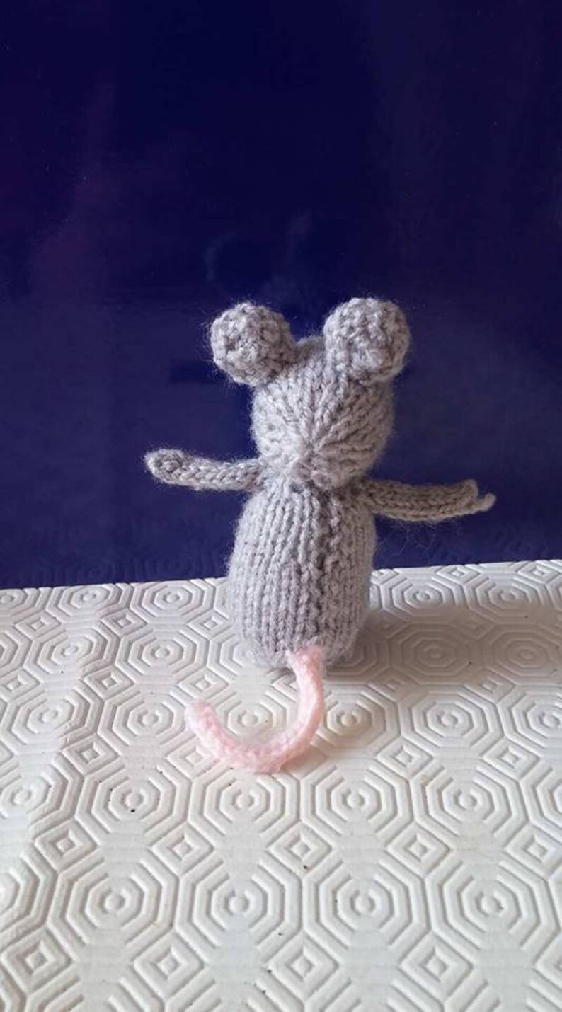 May include: A gray knitted mouse toy with pink ears and a pink tail. The mouse is sitting on a white surface with a hexagonal pattern.