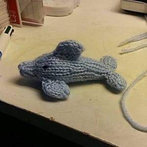 Puede incluir: Un juguete de peluche de delf&iacute;n de punto azul claro. El delf&iacute;n est&aacute; tumbado sobre una superficie blanca.