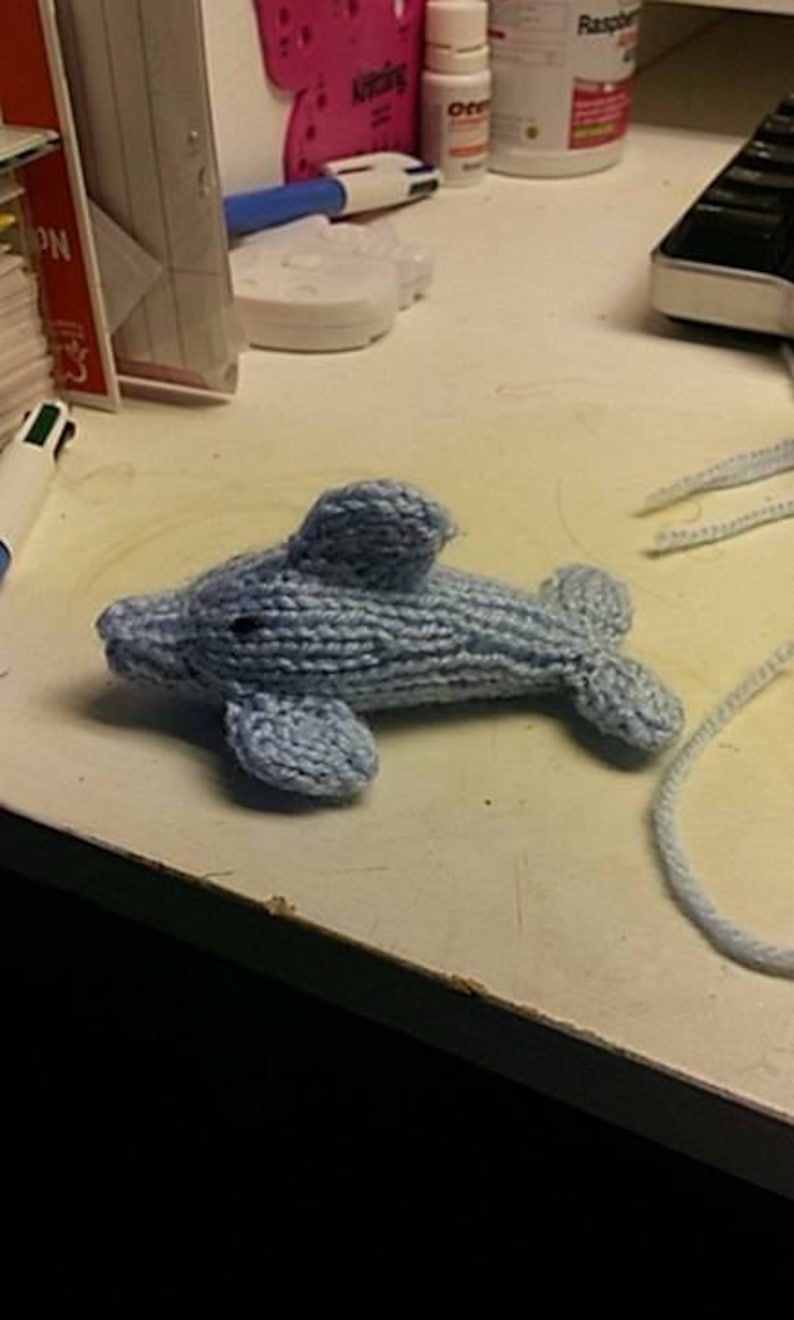 Puede incluir: Un juguete de peluche de delf&iacute;n de punto azul claro. El delf&iacute;n est&aacute; tumbado sobre una superficie blanca.