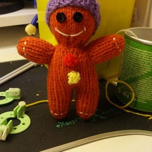 Puede incluir: Un juguete de jengibre tejido con un sombrero morado y un pompón blanco. El juguete tiene ojos negros, una sonrisa blanca y botones rojos y amarillos.