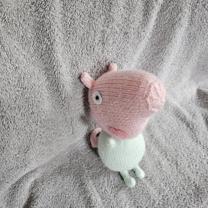 May include: A knitted pig toy in pink and light green. The pig has small ears, a snout, and a round body. The toy is placed on a grey, textured surface.