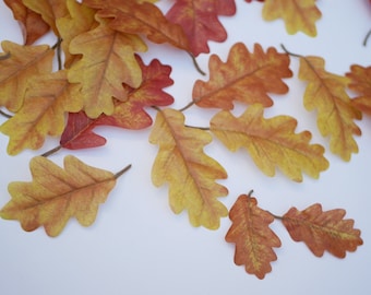 May include: Artificial oak leaves in shades of yellow, orange, and red are scattered on a white surface. The leaves have detailed veining and are arranged to suggest a natural fall scene, suitable for autumn decor.