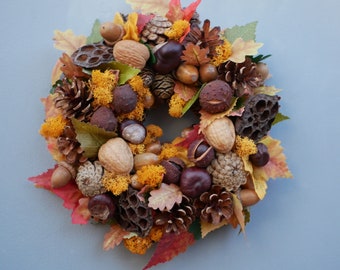 May include: A circular autumn wreath composed of natural elements. It features pine cones, walnuts, acorns, chestnuts, and dried seed pods, interspersed with colorful fall leaves and orange moss. The wreath is a decorative item.