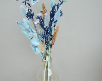 May include: A clear glass bottle holds a bouquet of dried flowers. The arrangement features blue and light blue dyed elements, including feathery plumes and seed heads. The flowers are set against a light gray background and sit on a wooden surface.