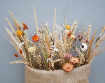 May include: A bouquet of dried flowers in a burlap sack. The arrangement includes wheat, orange, yellow, white, and pink flowers. The background is a solid light blue.