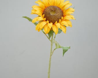 May include: A single artificial sunflower with bright yellow petals and a dark brown center. The flower sits in a silver vase against a gray background. The stem is green with a few green leaves.