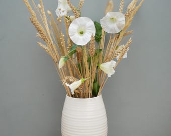 May include: A white ceramic vase with a ribbed design holds a bouquet of dried wheat stalks and white flowers. The wheat is a light brown color, and the flowers have delicate white petals with green centers. The vase is on a wooden surface.