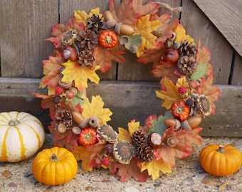 May include: A fall-themed wreath made of colorful autumn leaves, pinecones, acorns, and small pumpkins. The wreath is displayed against a wooden door. Three small pumpkins are placed in front of the wreath, and a white and yellow striped gourd is to the left.