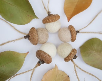 Può includere: Cinque decorazioni a forma di ghianda in feltro con cappucci marroni e cime in feltro bianco e beige. Le ghiande sono infilate con uno spago e disposte in cerchio su uno sfondo bianco.