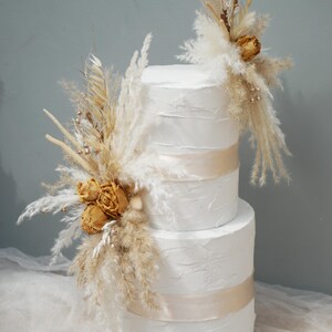 May include: A three-tiered white cake with textured frosting, decorated with dried flowers and pampas grass. The cake features a gold ribbon, creating an elegant, rustic design. Ideal for a wedding or special occasion.