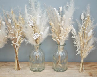 May include: Four small bouquets of dried pampas grass and white flowers in clear glass vases with a textured surface. The bouquets are arranged on a wooden surface.