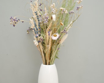 May include: A bouquet of dried flowers and grasses in a smooth, white, oval-shaped ceramic vase. The arrangement includes purple lavender, golden wheat stalks, white daisies, and green feathery foliage, creating a tall, textured botanical display.