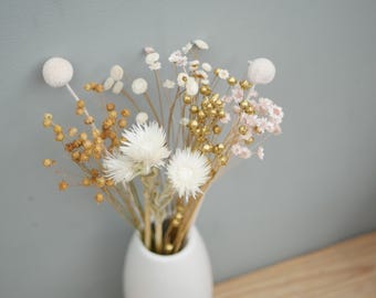 May include: A bouquet of dried flowers in a white ceramic vase, featuring white globe-shaped blossoms, small golden metallic spheres, light brown seed pods, and delicate white and pale pink filler flowers arranged on thin, slender stems.