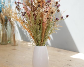 May include: A white ceramic vase containing a dried flower arrangement featuring various grasses, wheat stalks, and small purple, pink, and yellow blossoms. The bouquet displays a mix of textures and natural earth tones, standing upright in the smooth, oval-shaped container.