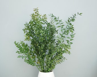 May include: A dense arrangement of artificial green ruscus branches featuring numerous small, pointed leaves in varying shades of olive and forest green. The stems are gathered together and placed inside a simple, solid white cylindrical container.