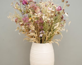 May include: A bouquet of dried flowers featuring pink roses, purple statice, and beige oat stalks arranged in a white ceramic vase with a ribbed, horizontal texture and a small square stamp on the lower front section of the vessel.