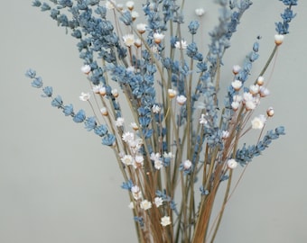 May include: A bouquet of dried lavender and small white flowers in a white vase. The lavender has a muted blue hue, and the arrangement is set against a neutral gray background. The stems are a light brown color.