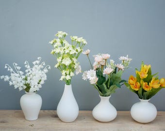 May include: Four white ceramic vases each hold a different arrangement of flowers. The flowers include white lily of the valley, yellow and white blooms, pink daisies, and yellow tulips. The vases are on a wooden surface.