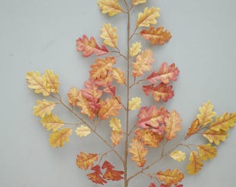 May include: A decorative artificial oak branch with leaves in shades of yellow, orange, and red. The branch has a brown stem and is set against a light blue-gray background. The leaves are detailed and textured, creating a realistic appearance.