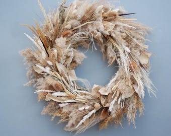 May include: A circular wreath composed of dried beige, tan, and white grasses, seed pods, and other natural elements. The wreath is displayed against a light blue background, creating a neutral and earthy aesthetic.