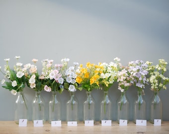 May include: A collection of colorful artificial flowers in clear glass bottles. The flowers include white, pink, yellow, and purple blooms. Each bottle has a small white tag with a number on it, arranged on a wooden surface.
