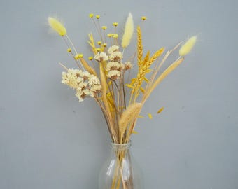 May include: A clear glass bottle holds a bouquet of dried flowers. The arrangement includes yellow, cream, and tan colored stems and blooms. The background is a solid gray wall, and the bottle sits on a wooden surface.
