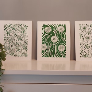 May include: Three framed prints with green floral designs. The prints feature different floral patterns, including clover, dandelions, and other flowers. The prints are all on white backgrounds.