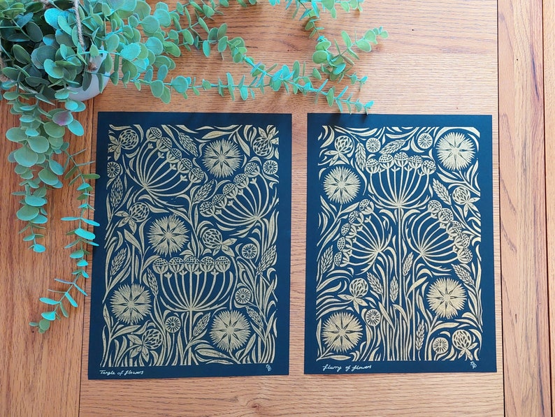 May include: Two floral art prints with gold and navy blue designs. The prints feature stylized flowers and foliage. One print is labeled "Temple of Flowers" and the other is labeled "Flowery of Flowers". The prints are displayed on a wooden surface.