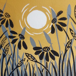 May include: A linocut print of a stylized sun with a white circle and rays, set against a yellow background. Black and gray stylized flowers and grass are in the foreground.