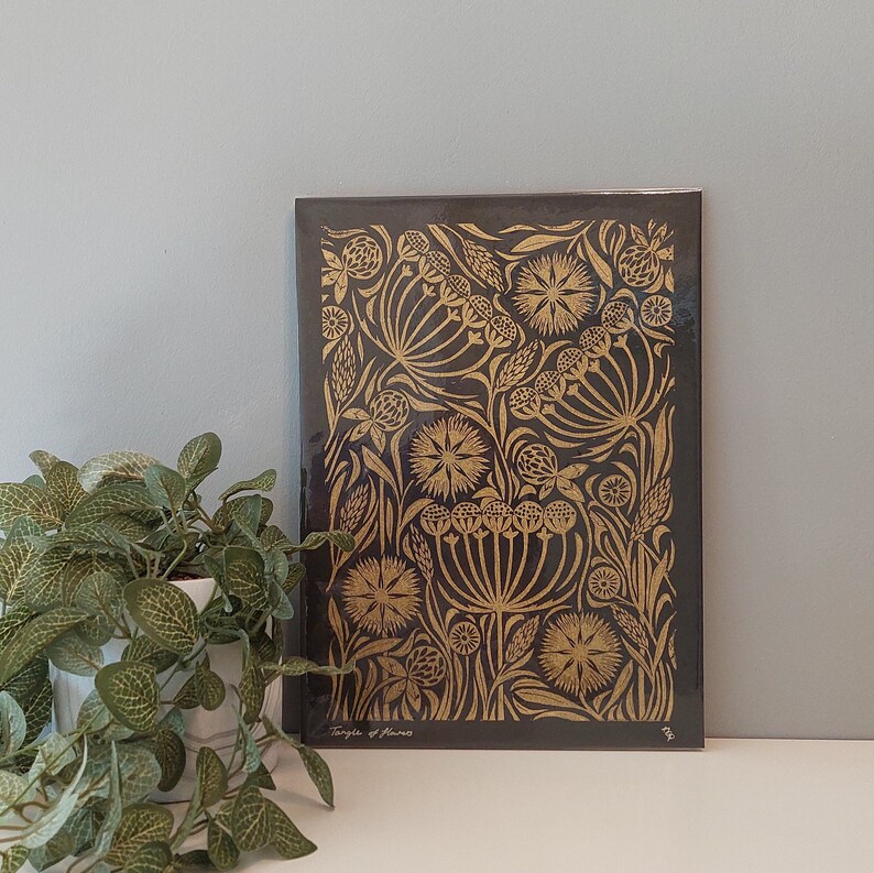 May include: A black journal with a gold floral design. The cover features an intricate pattern of stylized flowers and foliage. The words "Tangle of Flowers" are printed in the lower left corner. A potted plant is in the foreground.