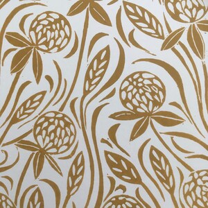 May include: A white background with a repeating pattern of gold clover flowers and leaves. The clover flowers are stylized and have a textured look.
