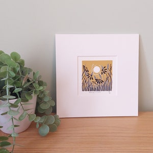 May include: A framed print of a stylized illustration of a field of flowers with a white sun in the background. The flowers are blue and black on a yellow background. The print is matted in white and has a white frame.