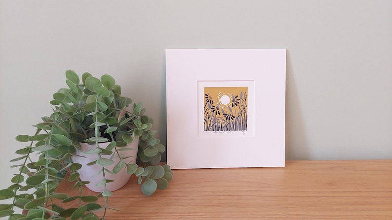 May include: A framed print of a stylized illustration of a field of flowers with a white sun in the background. The flowers are blue and black on a yellow background. The print is matted in white and has a white frame.
