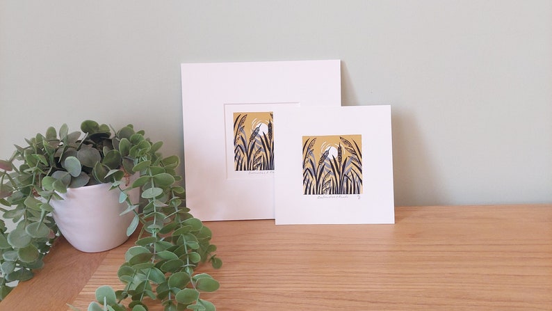 May include: Two framed prints of a stylised illustration of tall grass with a white bird in the centre. The background is a yellow-gold colour. The prints are in white frames and are on a wooden surface.