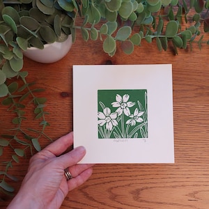 May include: A white card with a green border and a white print of daffodils on a green background. The card says "Daffodils" at the bottom.