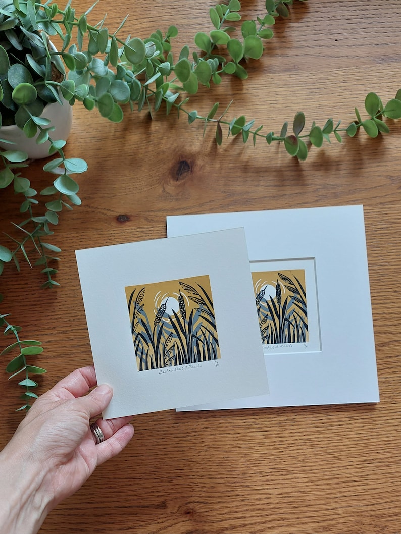 May include: A framed print of a nature scene with tall grasses and a yellow moon in the background. The print is titled "Cattails & Reeds".