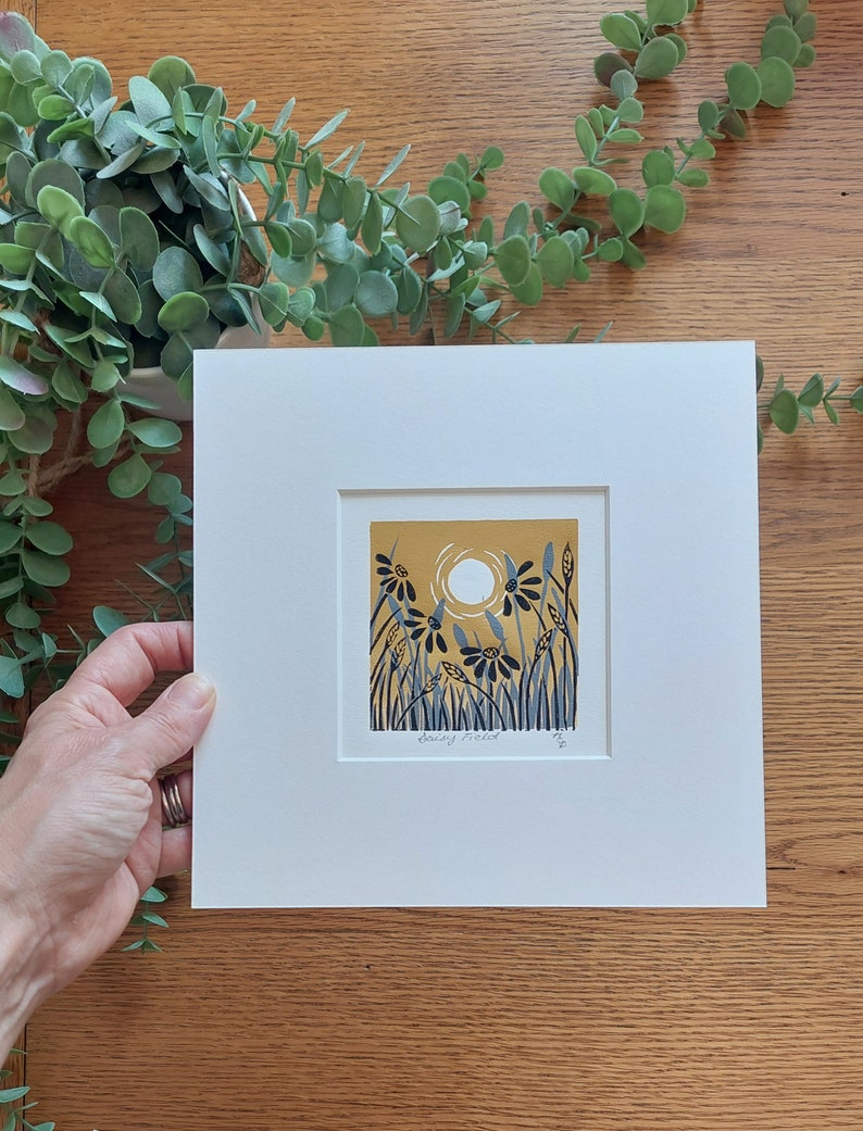 May include: A framed print of a yellow and blue illustration of a field of flowers with a sun in the background. The print is signed "Daisy Field" and numbered 20.