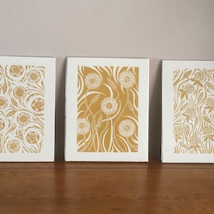 May include: Three framed prints with a white background and gold floral designs. The prints feature different floral patterns, including a repeating pattern of flowers and leaves, a pattern of stylized flowers with long stems, and a pattern of clover-like flowers.